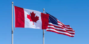 "علما الولايات المتحدة الأمريكية وكندا يرفرفان معًا تحت سماء زرقاء صافية، يرمزان إلى العلاقات الثنائية بين البلدين."